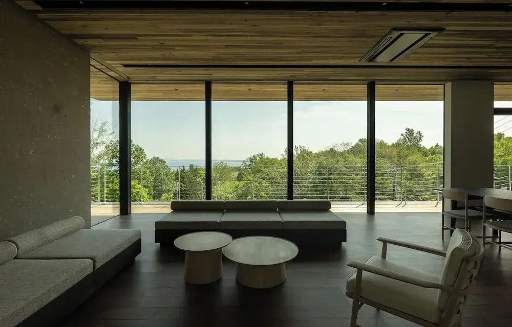 Modern living room with large windows overlooking a lush forest and distant water view. Minimalist furniture includes sofas and round coffee tables.
