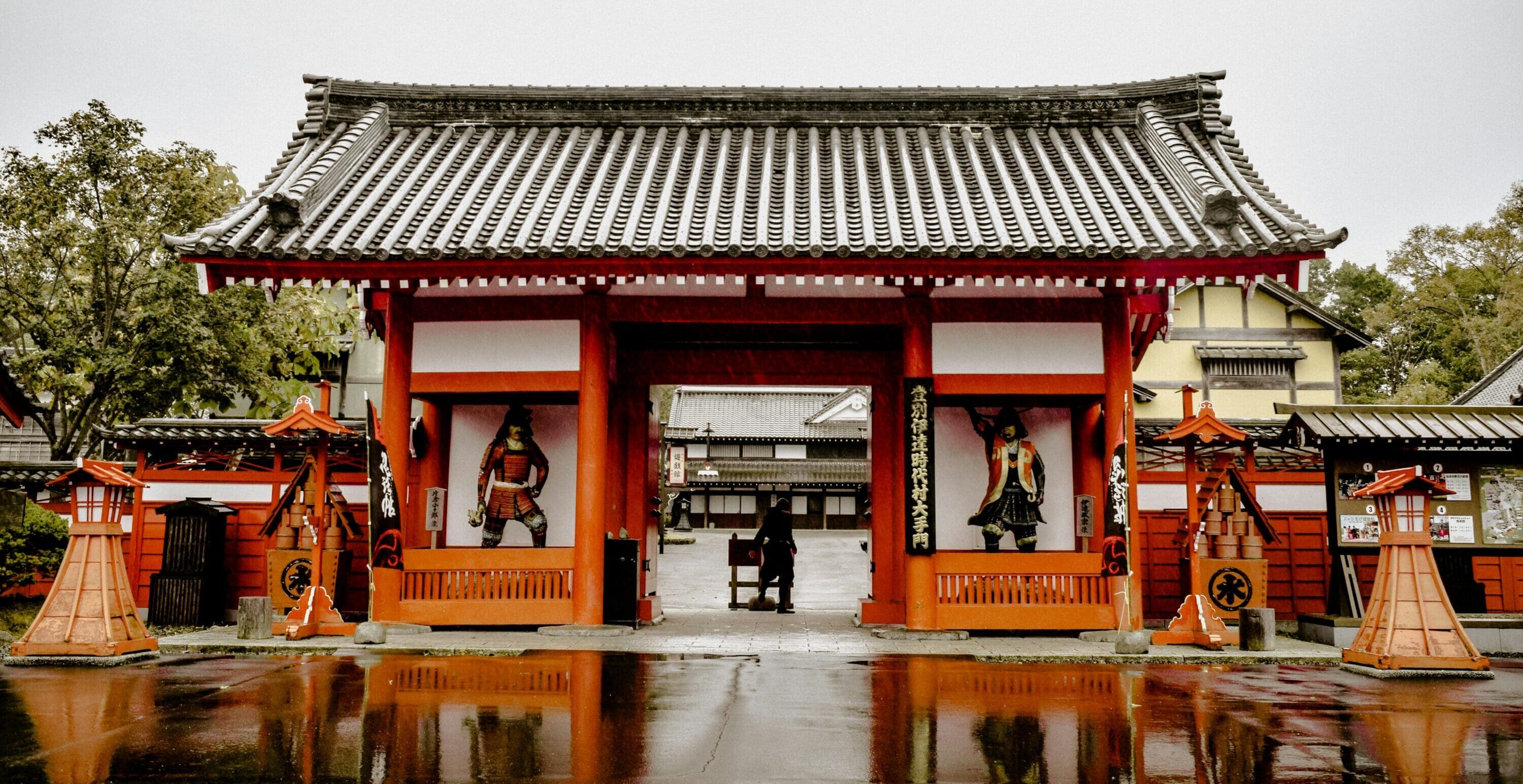 A traditional Japanese temple gate with vibrant red wooden pillars and a black-tiled roof, reflected in rain-soaked ground, creating a serene atmosphere.