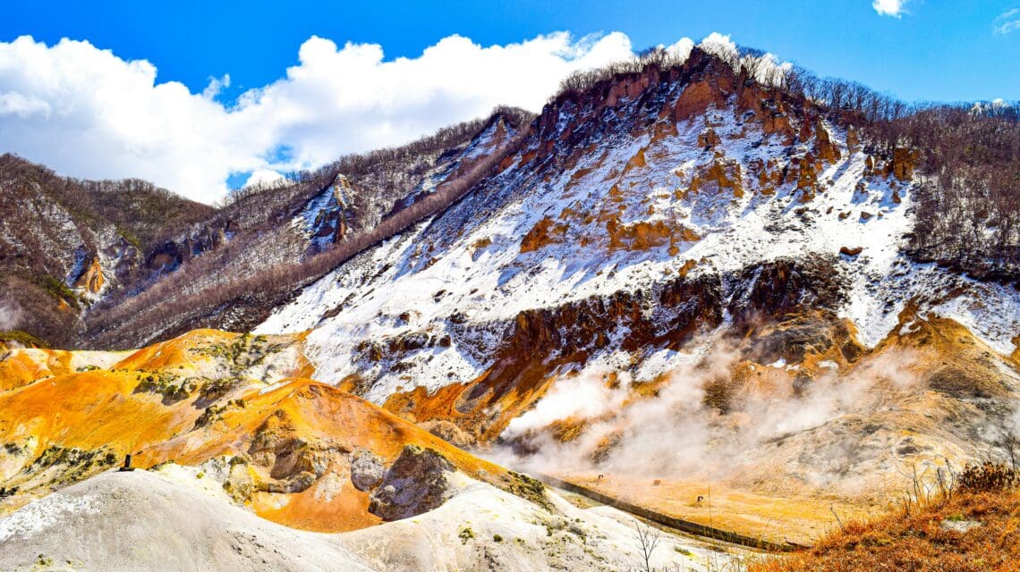 Snow-dusted volcanic landscape with orange earth and rising steam, set against a bright blue sky and scattered clouds, creating a rugged, natural beauty.