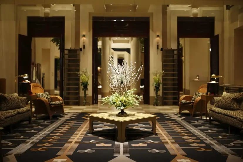 Elegant hotel lobby with warm lighting, featuring a central flower arrangement on a square table. Plush sofas and chairs line patterned carpet. Luxurious ambiance.