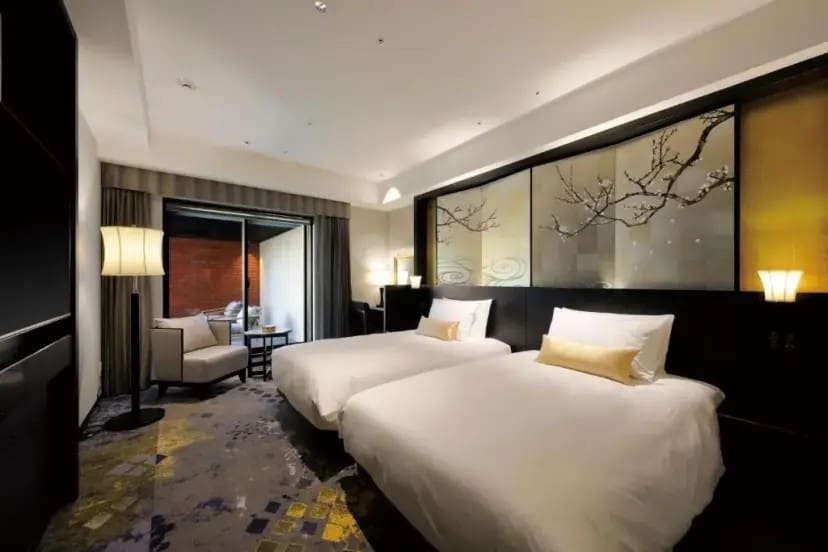 Elegant hotel room with two neatly made twin beds, light wood accents, and decorative wall panels with tree branch designs. Soft lighting and a cozy seating area by a window to the left create a calm, inviting atmosphere.