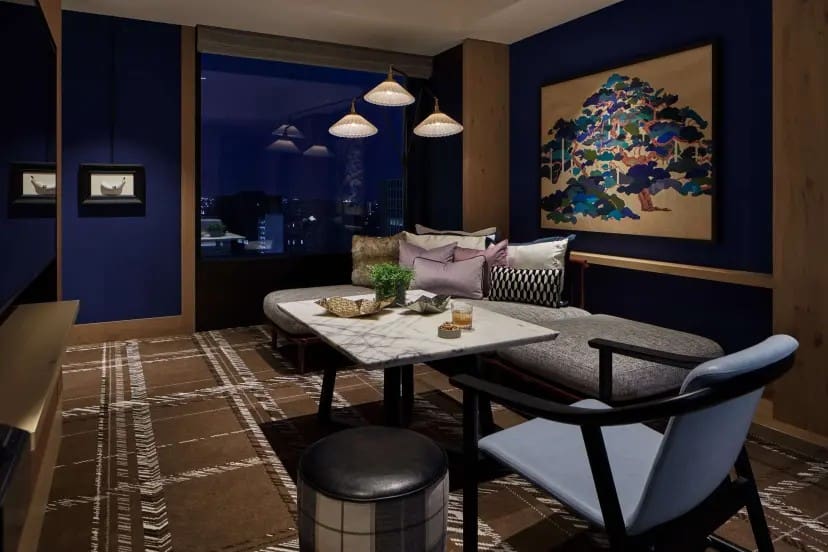 Cozy lounge with deep blue walls, featuring a marble coffee table, plush couch with pillows, artistic wall painting, and warm pendant lights.