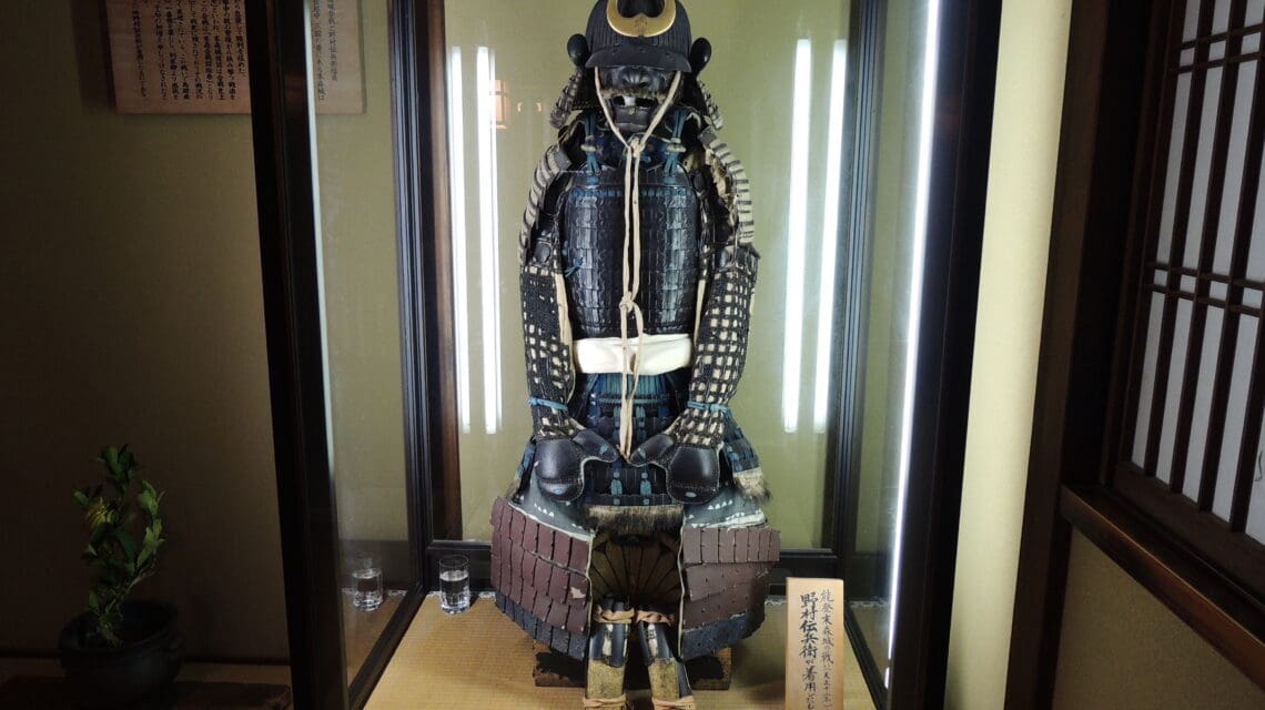 Samurai armor displayed in a glass case, featuring intricate blue and black patterns and a crescent moon crest. The room has a traditional Japanese aesthetic.