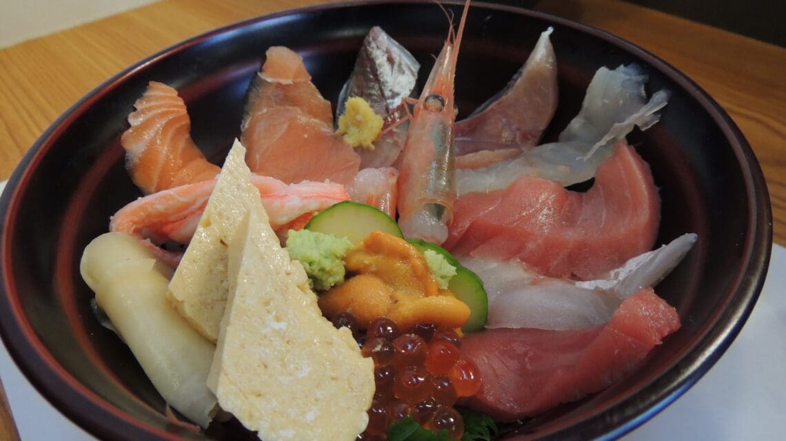 A black bowl with assorted sashimi, including salmon, tuna, shrimp, and ikura, beautifully arranged on rice. Topped with wasabi and garnishes.