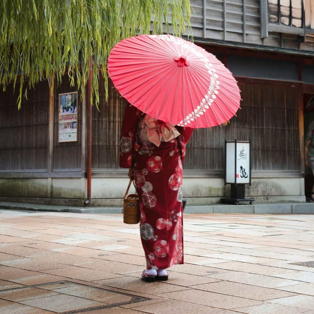 A person in a traditional red kimono with floral patterns holds a large red parasol, walking past wooden buildings under a lush green willow tree.