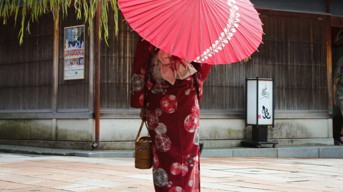 A person in a traditional red kimono with floral patterns holds a large red parasol, walking past wooden buildings under a lush green willow tree.