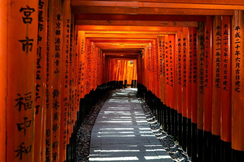 A sunlit path winds through vibrant orange torii gates lined with Japanese inscriptions at Fushimi Inari Shrine, evoking a sense of tranquility and cultural richness.