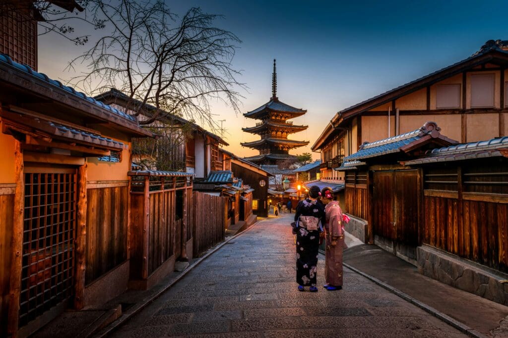 Two people in traditional kimonos stand on a narrow street in Kyoto at dusk, with wooden houses lining the path and a pagoda tower in the background.