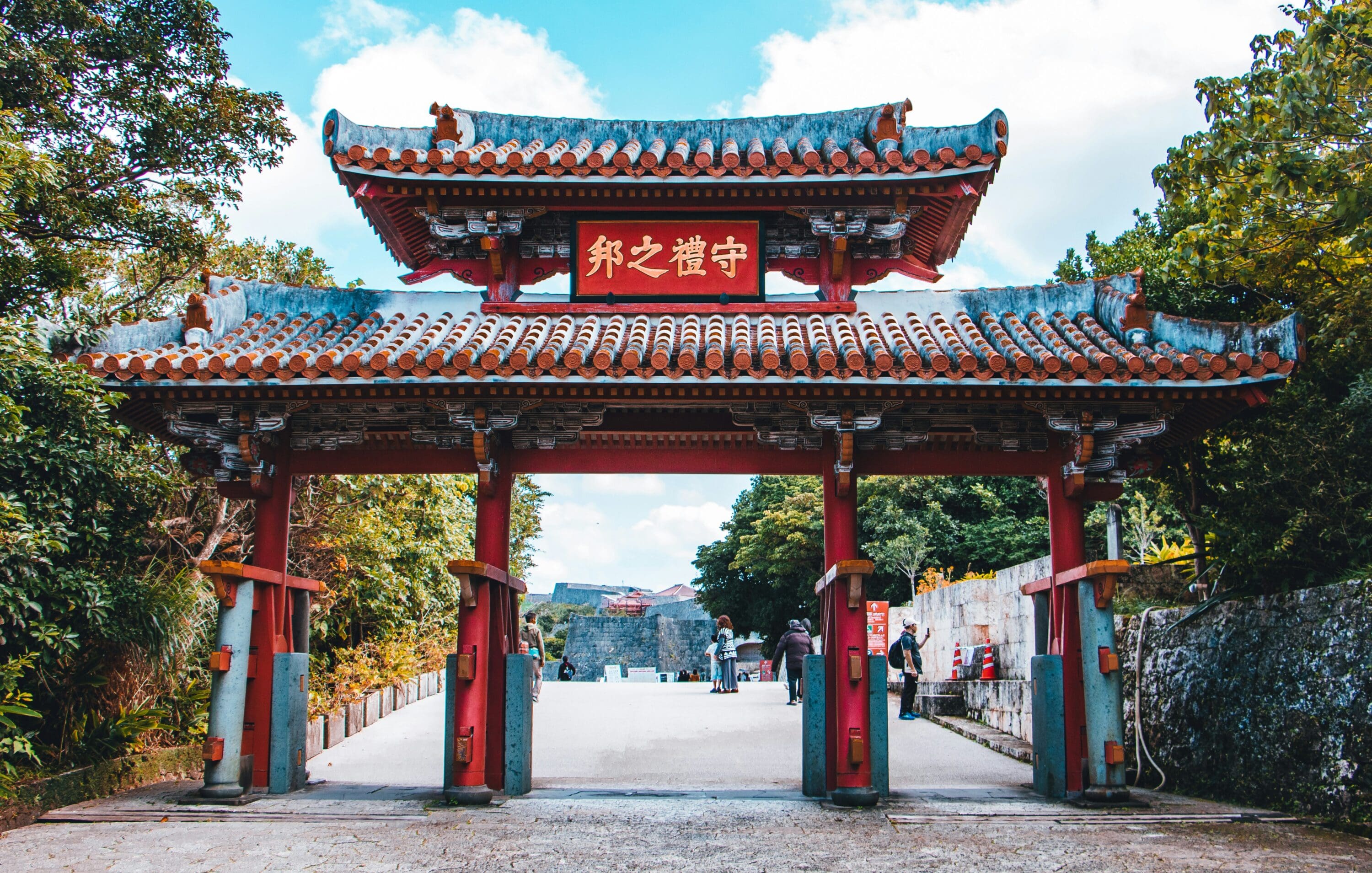 Traditional red and gray Japanese gate with a decorative tiled roof, set in a lush green environment. People walk through, creating a serene atmosphere.