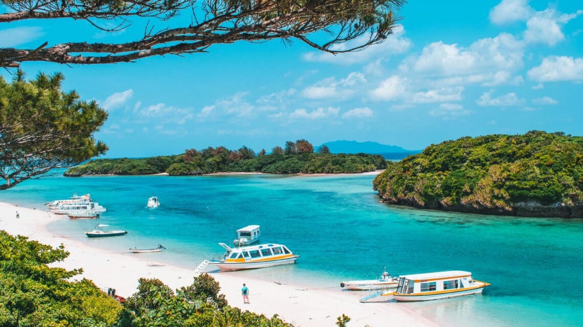 Tropical beach scene with clear turquoise water, white sand, and anchored boats. Lush green islands and a bright blue sky complete the serene landscape.