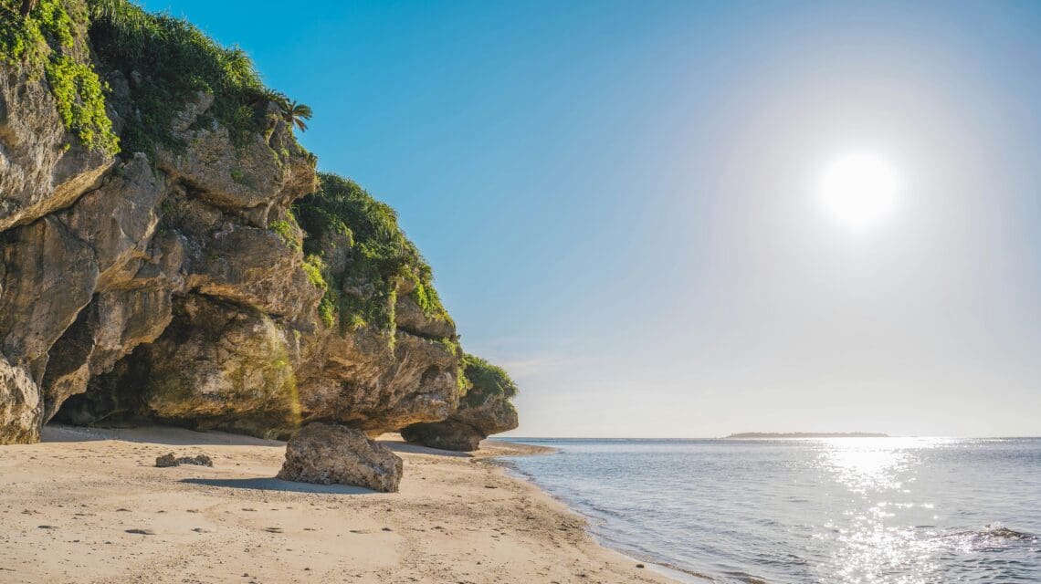 Sunlit beach with large rocky cliffs covered in green foliage on the left, and calm blue ocean on the right under a clear sky, conveying tranquility.
