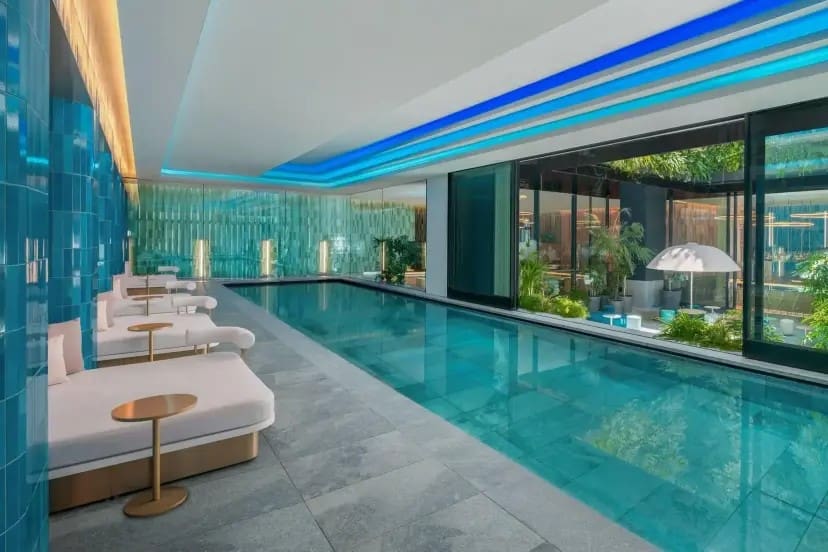 Luxurious indoor pool area with turquoise tiles, plush white loungers, and sleek gold tables. Large windows reveal lush greenery and outdoor seating.