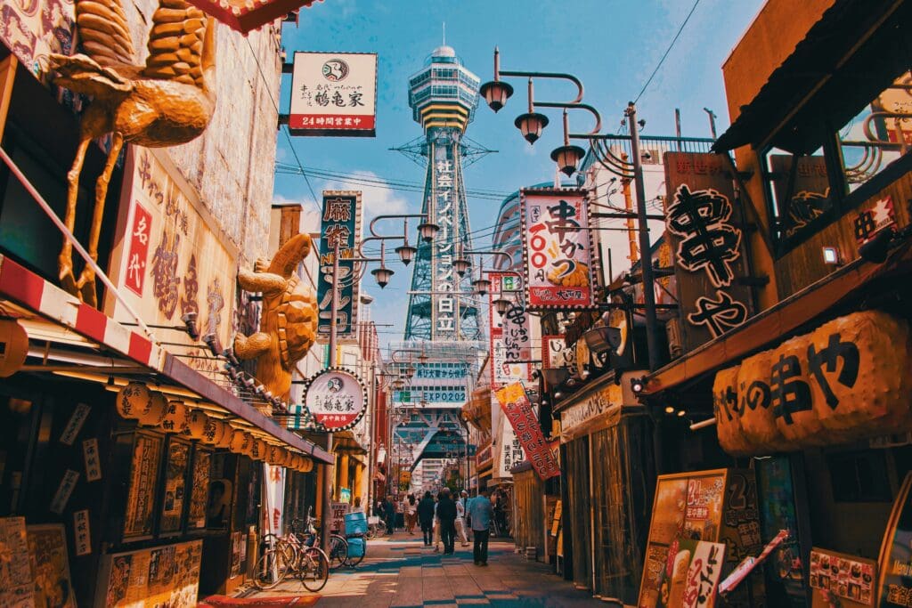 Bustling Japanese street scene with colorful signs, large food sculptures, and Tsutenkaku Tower in the background under a bright blue sky.