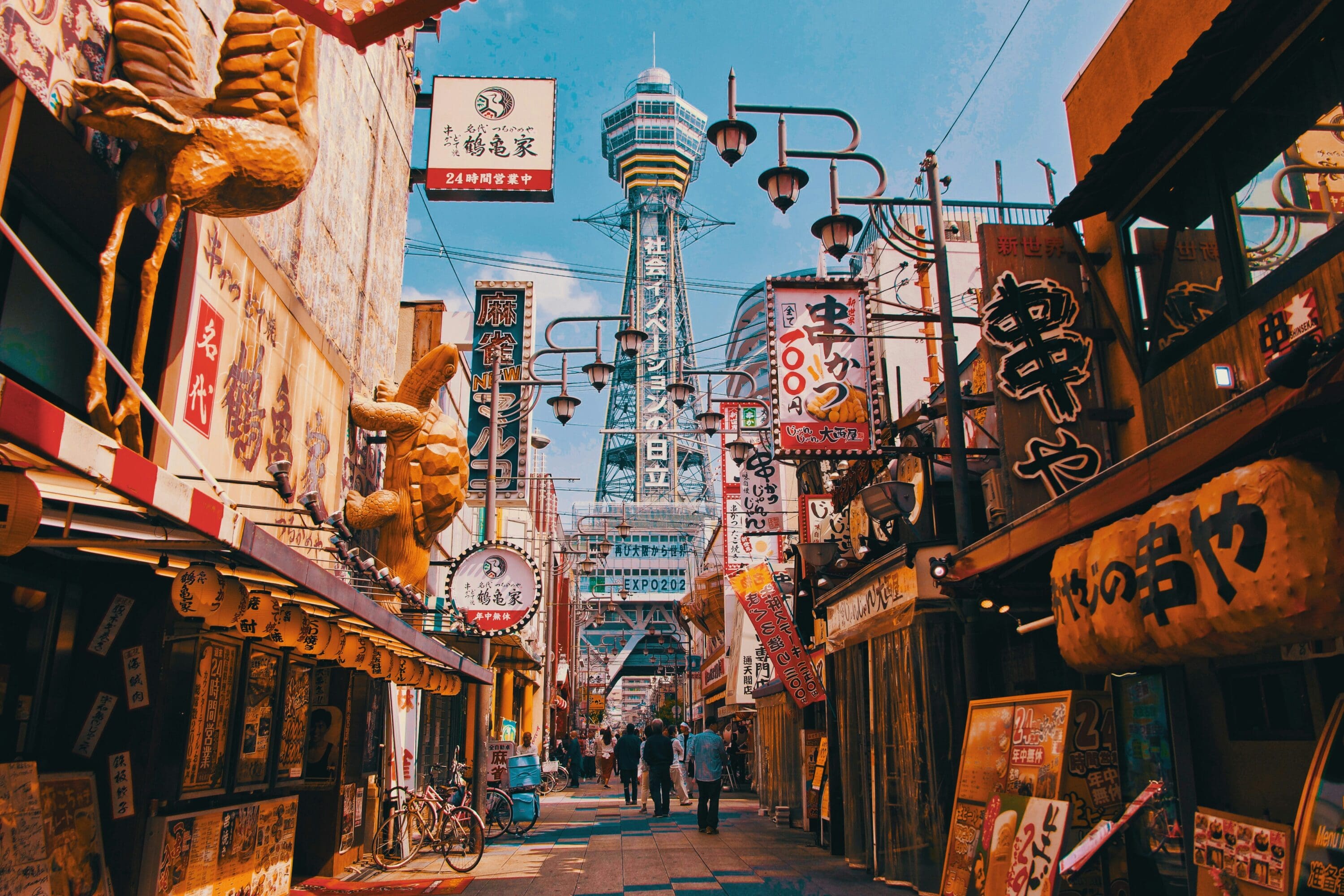 Bustling Japanese street scene with colorful signs, large food sculptures, and Tsutenkaku Tower in the background under a bright blue sky.