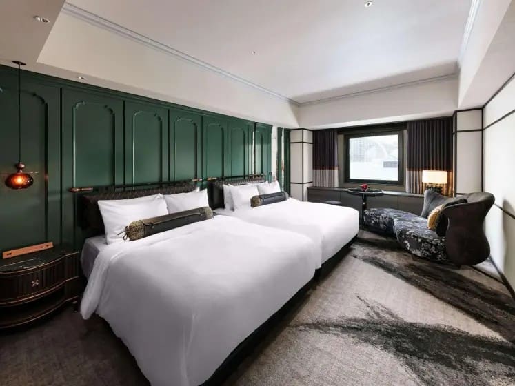 Modern hotel room with two neatly made beds, green accent wall, and stylish furnishings, including a sofa by a window. The ambiance is elegant and inviting.