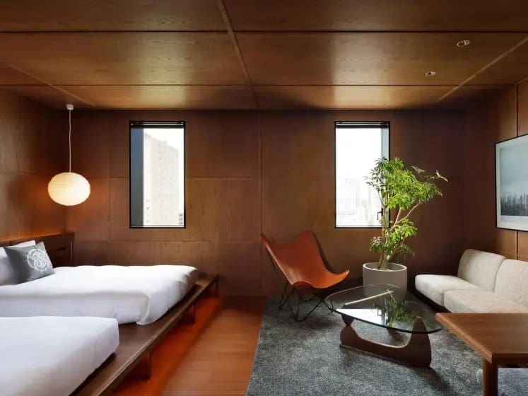 Modern minimalist bedroom with wooden walls, featuring a low platform bed, butterfly chair, glass coffee table, potted plant, and ambient lighting.