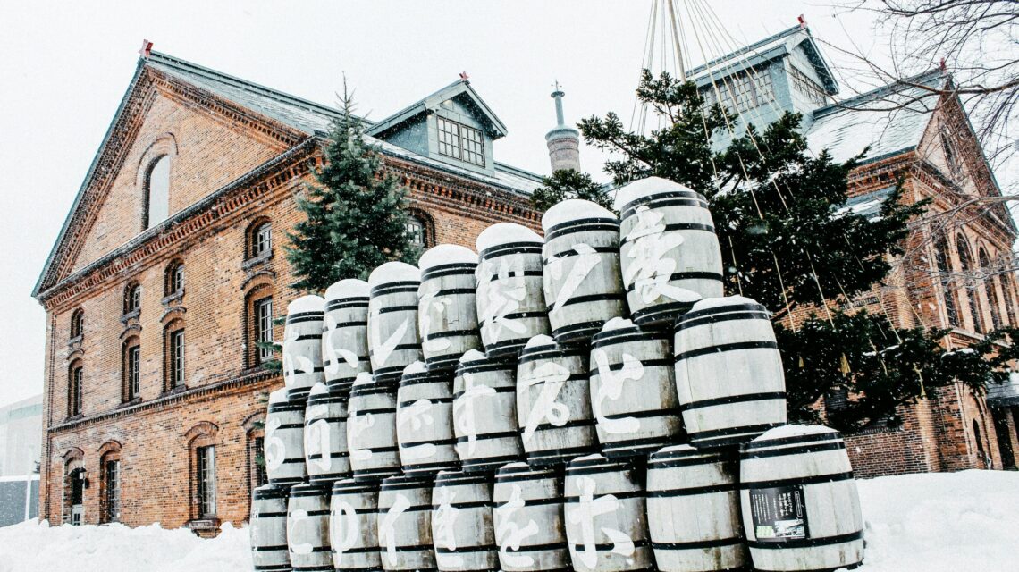 Snowy scene of a brick brewery building with a stack of white barrels in front, adorned with Japanese characters. Evergreen trees add greenery. Tranquil winter atmosphere.