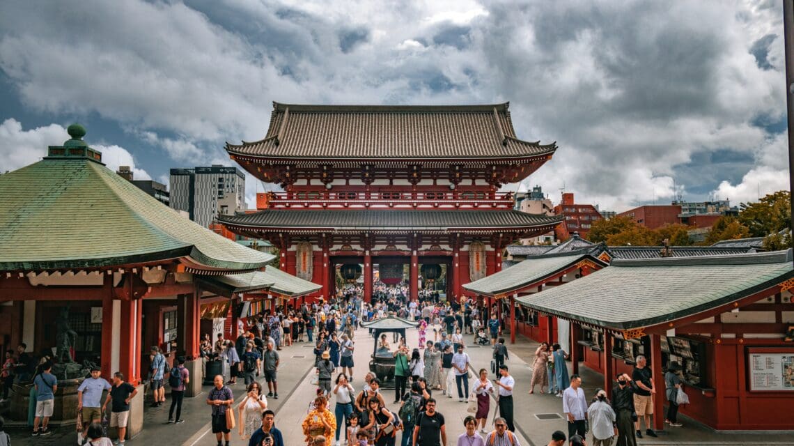 Crowds gather at a traditional Japanese temple with ornate red and green roofs under a cloudy sky, conveying a vibrant, bustling atmosphere.