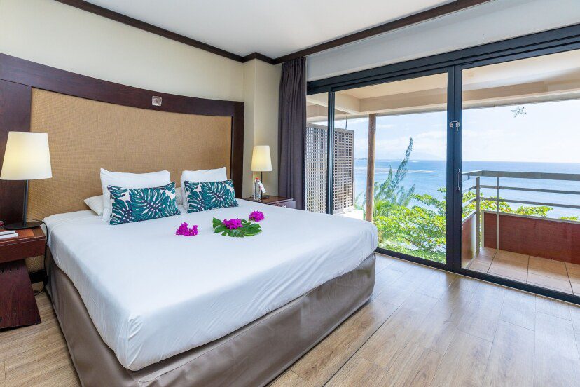 Modern hotel room with a large bed, tropical-themed pillows, and purple flowers. Glass doors open to a balcony with a scenic ocean view. Bright and inviting ambiance.