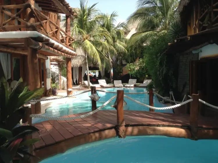 Tropical resort pool with wooden bridge, surrounded by palm trees and lounge chairs. Rustic architecture creates a serene and inviting atmosphere.