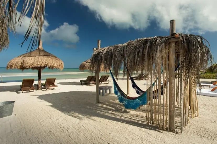 Tranquil beach scene with hammocks under a thatched roof, lounge chairs shaded by umbrellas, sandy shore, turquoise sea, and a partly cloudy sky.