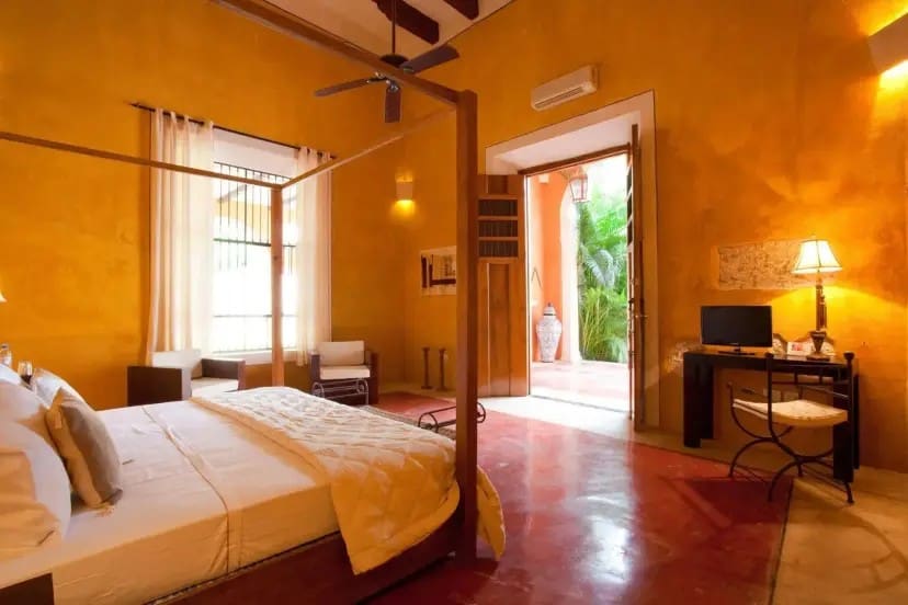 Cozy bedroom with orange walls, a canopy bed, and white bedding. Sunlight streams through large windows and an open door reveals lush greenery.
