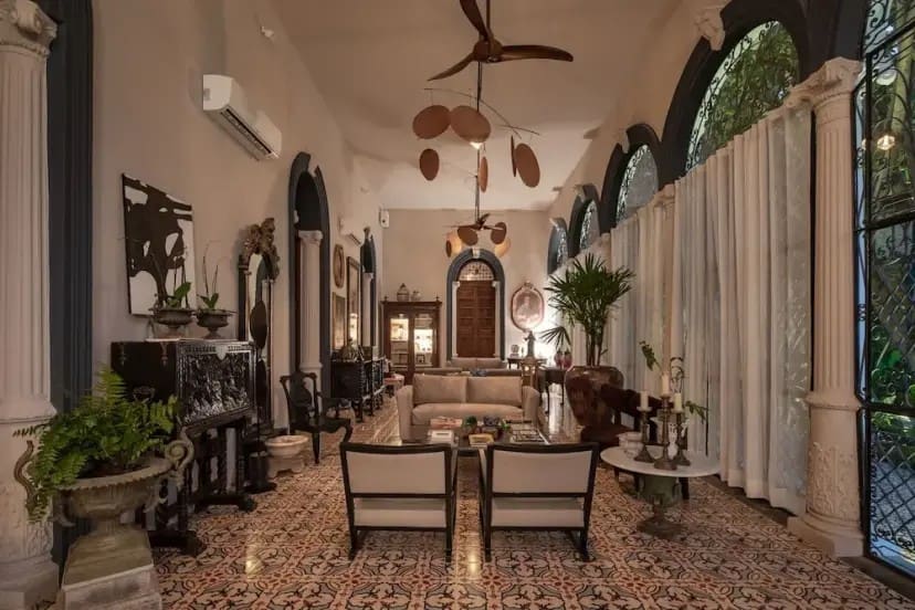 Elegant living room with high ceilings, ornate tiled floor, and arched windows draped in curtains. Central seating area with chairs and sofa, surrounded by lush plants and classic decor. Warm, inviting atmosphere.