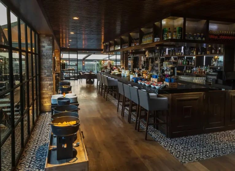 Modern restaurant interior with warm lighting, featuring a long wooden bar with high chairs and shelves stocked with bottles. Bright, inviting atmosphere.