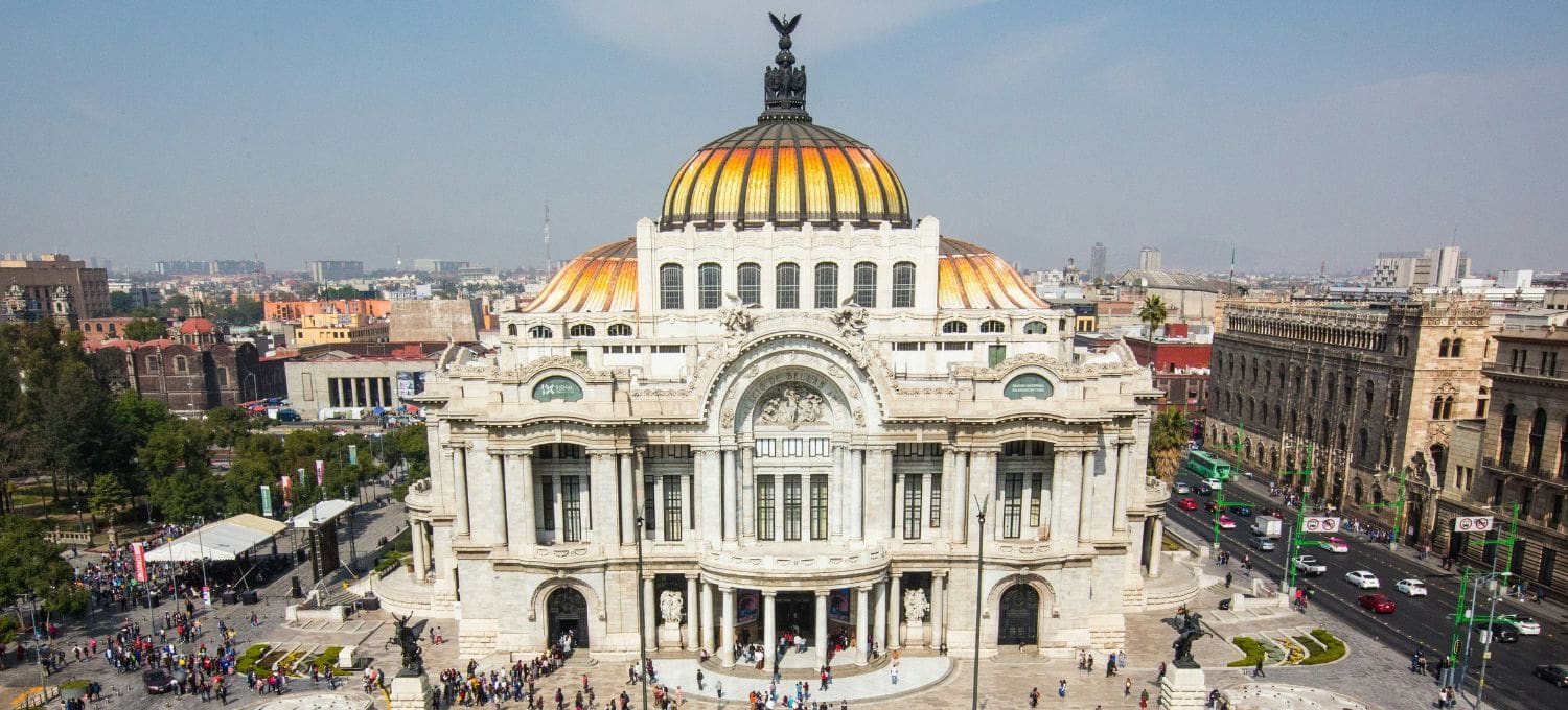 Aerial view of a grand, ornate building with a yellow-orange domed roof, crowded with people, set in a bustling cityscape under a clear blue sky.