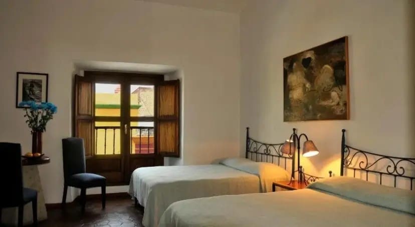 Cozy room with two single beds and ornate black metal headboards, lit warmly by a lamp. A window with wooden shutters, art, and flowers enhance the serene atmosphere.