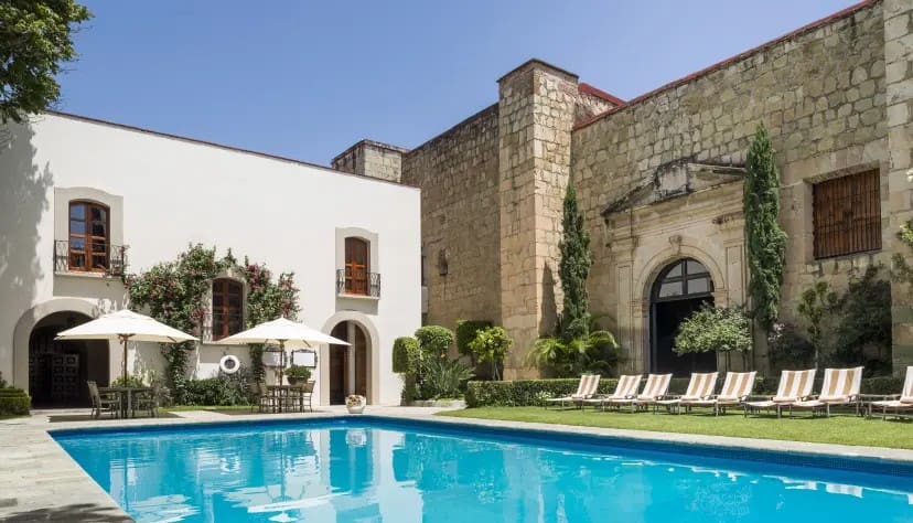 A serene courtyard with a bright blue pool surrounded by a historic stone wall and a white building. Lounge chairs and umbrellas create a relaxing atmosphere.