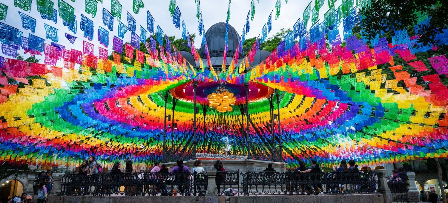 A vibrant ceiling of colorful papel picado banners forms a circular pattern over a gazebo. People below admire the festive, kaleidoscopic display.