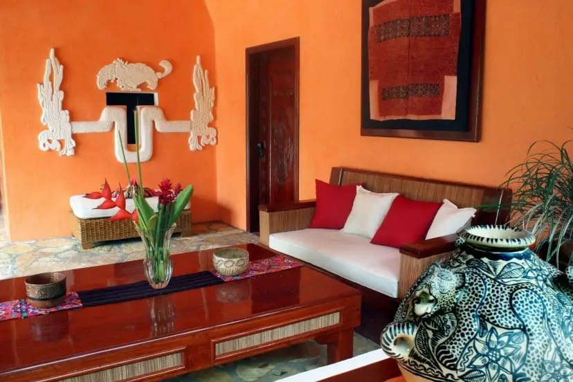 Inviting room with orange walls, a wooden couch with red and white cushions, a polished table, floral centerpiece, and tribal-style decor exuding warmth.