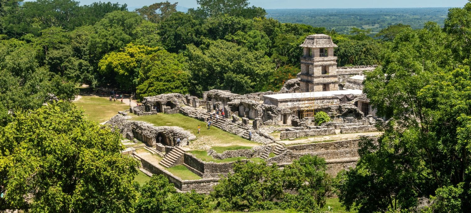Ancient stone structure at Palenque, Mexico, surrounded by lush greenery. The scene is serene and historical, with a few visitors exploring.
