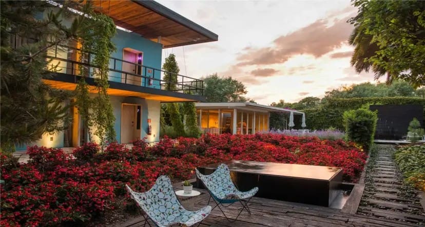 Modern house with lush garden, featuring vibrant red flowers and green vines. Two patterned chairs face a sleek water feature, under a sunset sky.