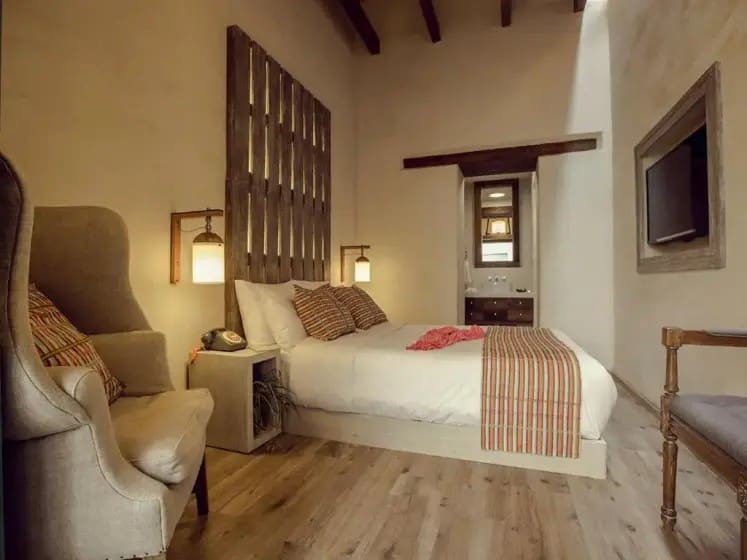 Cozy bedroom with wooden accents features a bed adorned with striped pillows and a blanket. Warm lighting, an armchair, wall-mounted TV, and wooden beams add rustic charm.