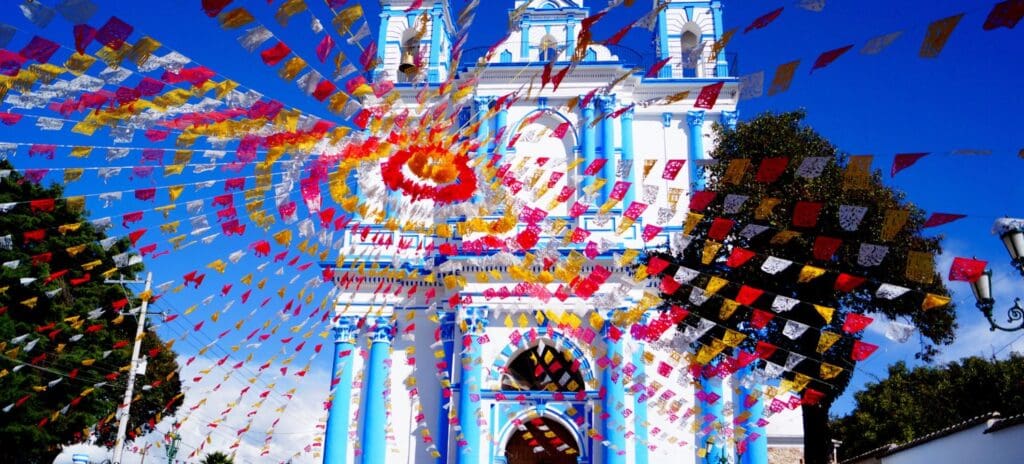 Colorful paper flags radiate from a vibrant church with blue accents against a clear sky, creating a festive and joyful atmosphere.