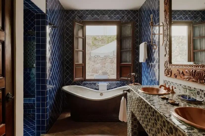 Luxurious bathroom with diamond-patterned blue tiles, a vintage freestanding tub, large wooden-framed window, and ornate mirror above dual copper sinks.