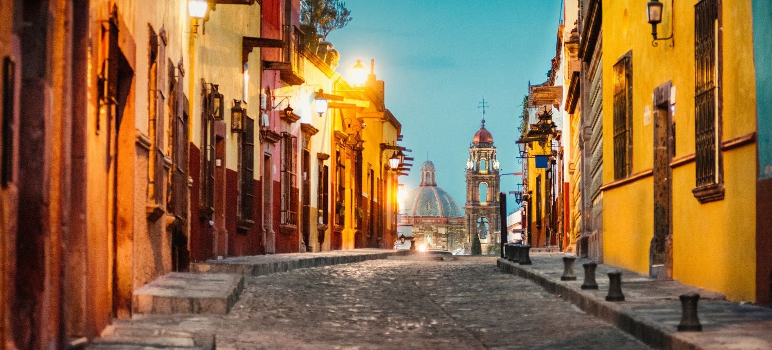 Narrow cobblestone street flanked by colorful buildings with warm lights, leading to a distant illuminated church dome and bell tower under a twilight sky.