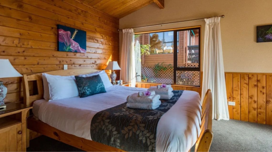 Cozy wooden bedroom with a large bed, white and teal bedding, and folded towels. Sunlight streams through a window with white curtains, creating a warm, inviting atmosphere.