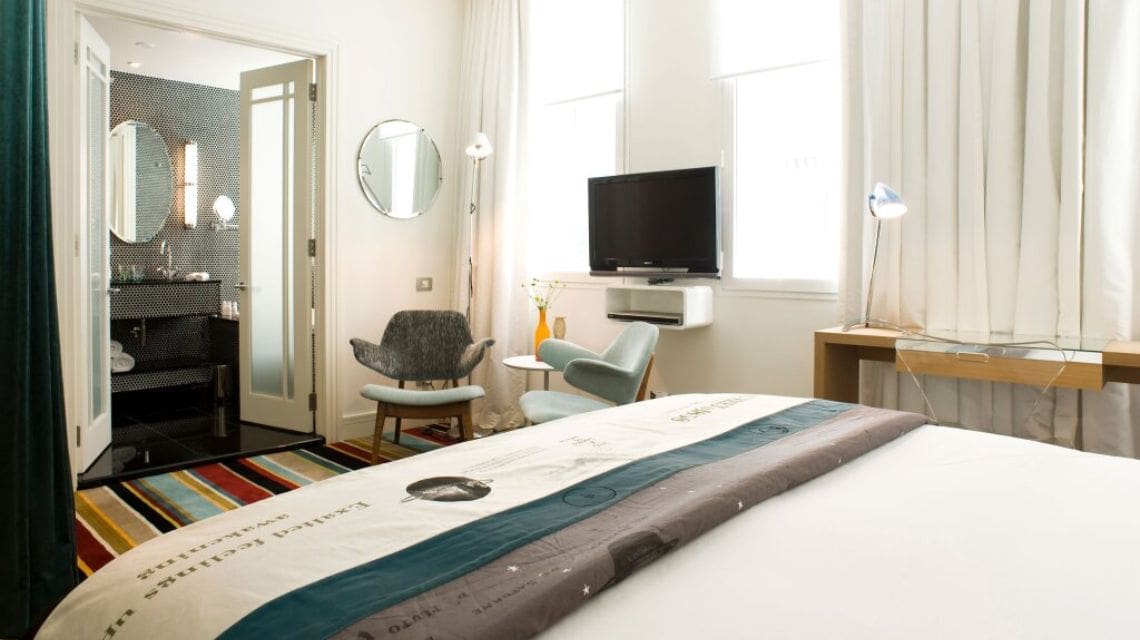 Interior shot of a hotel room with a queen size bed, bright light coming in through a window