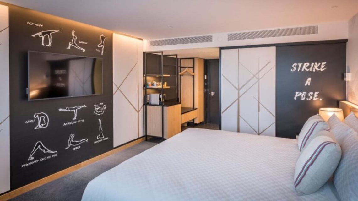 Modern hotel room featuring a bed with striped pillows, yoga pose illustrations on the wall, and "Strike a Pose" text. Warm lighting creates a cozy atmosphere.