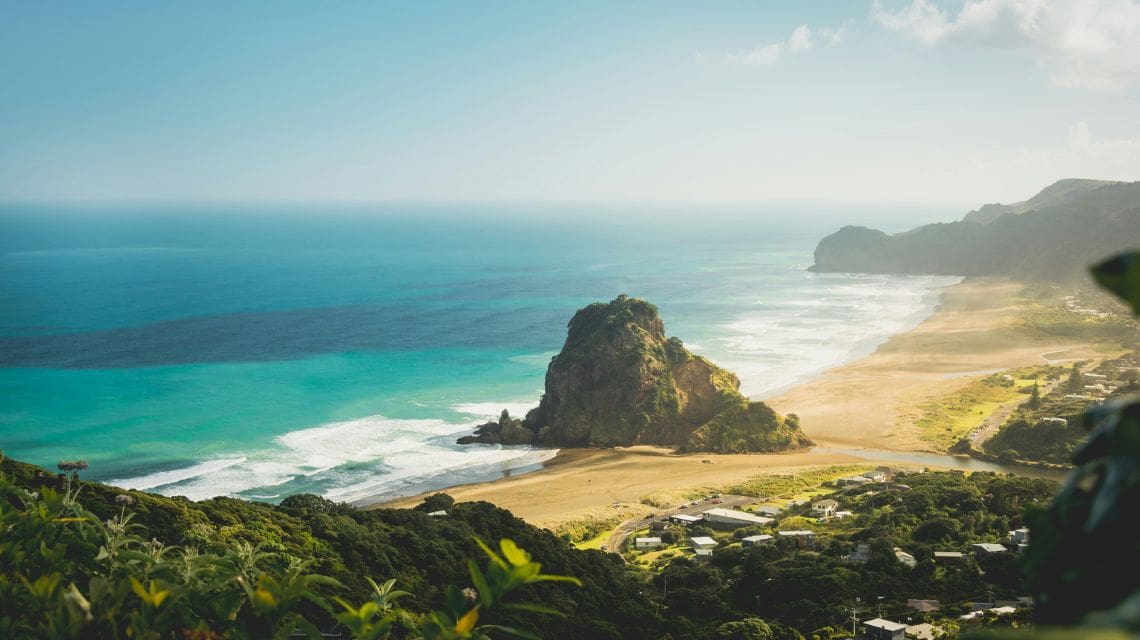 Aerial view of a scenic beach with turquoise waves and rugged cliffs. A large rock formation on the sandy shore is surrounded by lush greenery and calm dwellings.