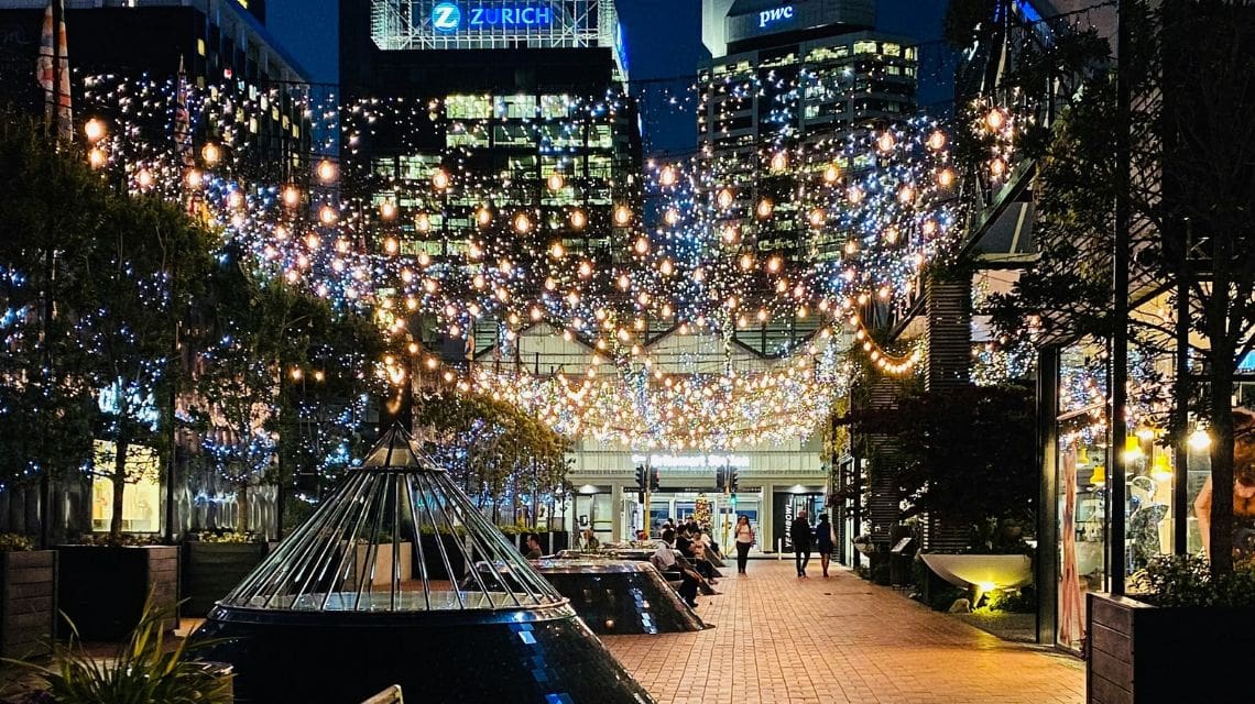 A vibrant urban alleyway lit by strings of colorful fairy lights, creating a cozy, festive atmosphere. People stroll leisurely beneath the twinkling canopy.