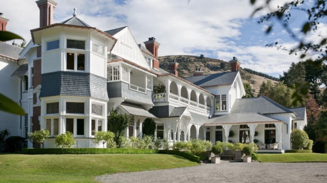Victorian-style mansion with white siding and red brick chimneys, surrounded by manicured greenery. Set against rolling hills under a blue sky.