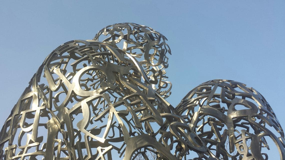 A sculpture of human figures made of interwoven metal letters against a clear blue sky. The artwork conveys a sense of unity and modernity.