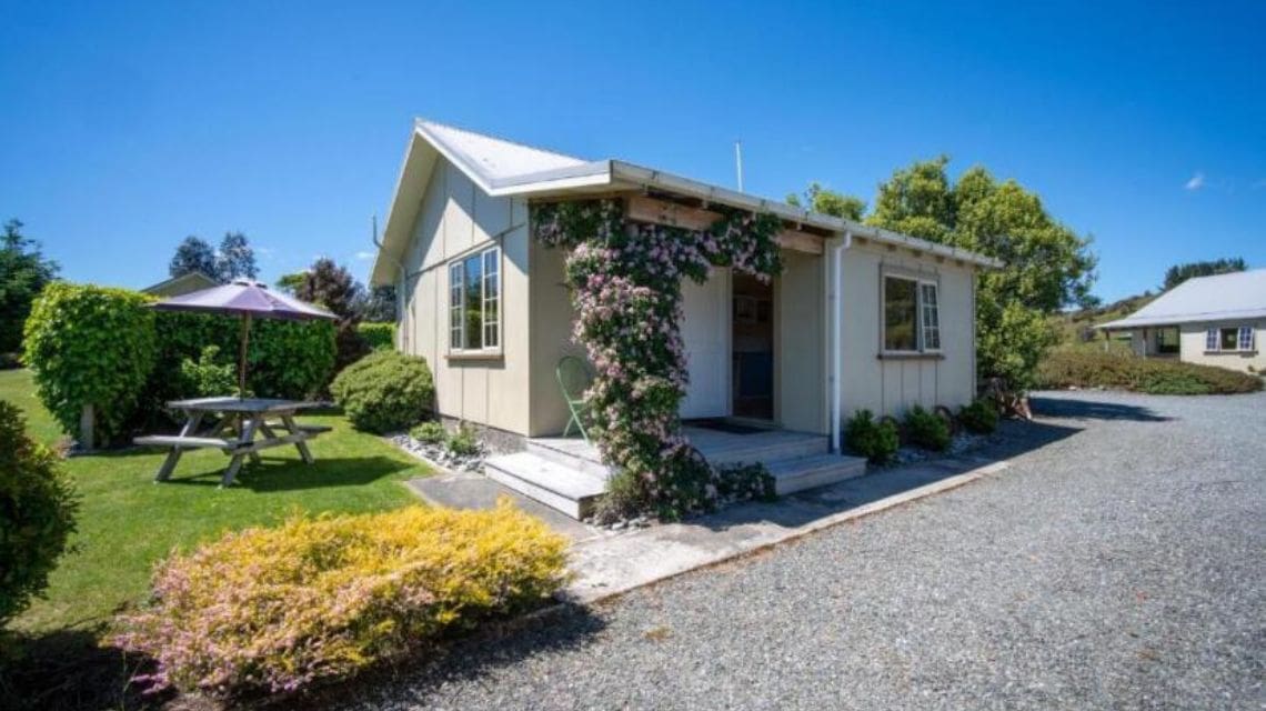 Small cottage with ivy on the porch posts, surrounded by a lush garden. A picnic table with an umbrella sits nearby. Bright, sunny day with clear skies.