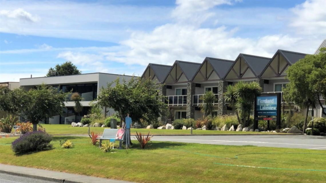 Modern motel with a series of pointed roofs, surrounded by neat lawns and trees under a partly cloudy sky.