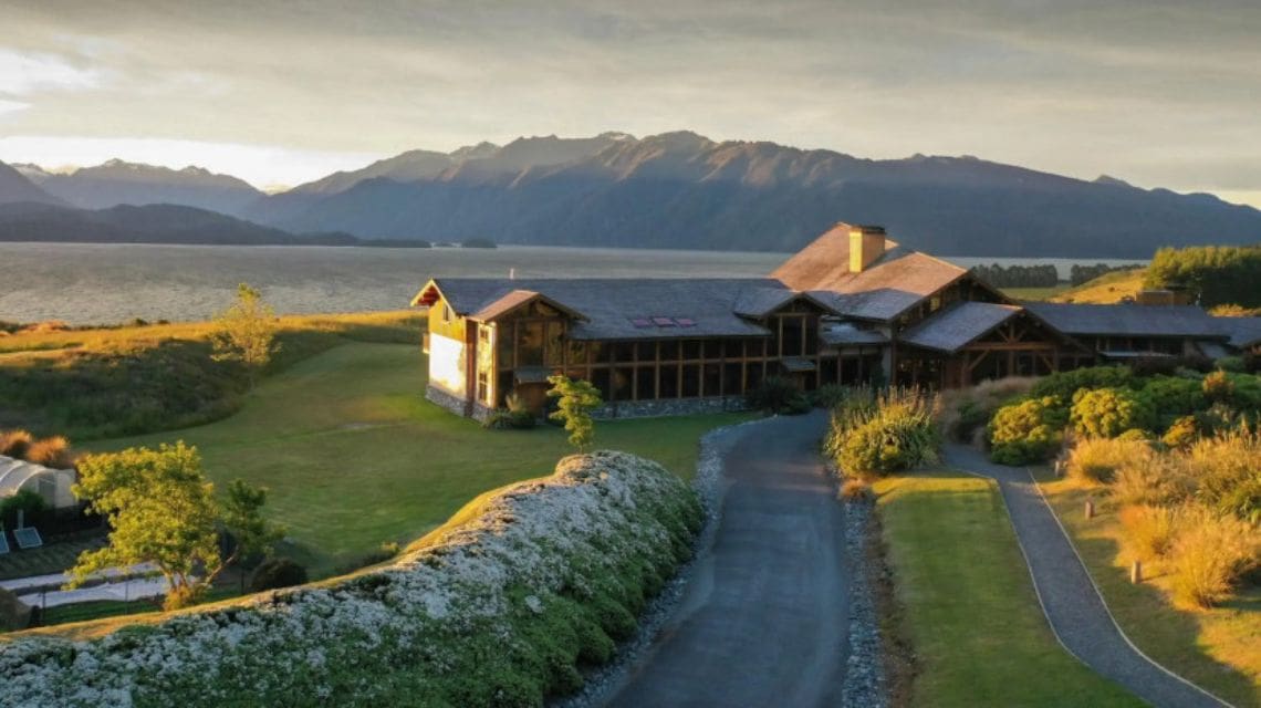 A large lakeside lodge with wooden architecture is surrounded by lush greenery and gardens. Mountains rise in the background under a clear sky.