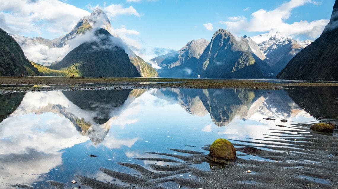 Snow-capped mountains and lush greenery reflect in a serene lake under a bright blue sky, evoking tranquility and natural beauty.